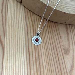 May include: A silver necklace featuring a circular pendant with a hammered texture. The pendant has a central square frame containing a small, round, red stone. The necklace is suspended from a thin, delicate silver chain with a box link design.
