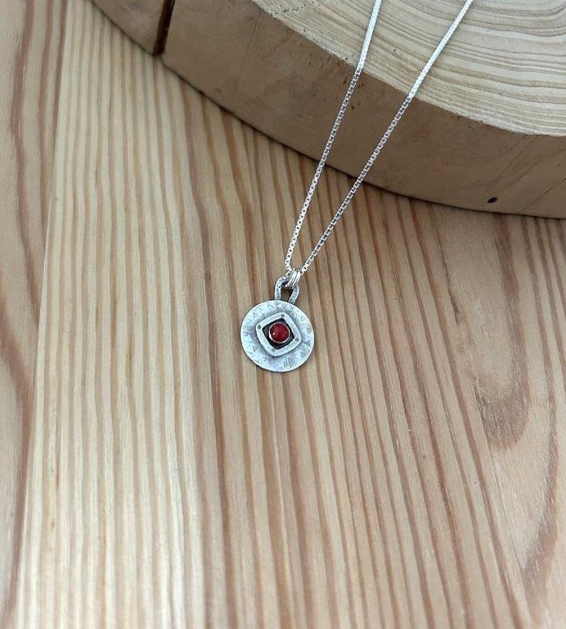 May include: A silver necklace featuring a circular pendant with a hammered texture. The pendant has a central square frame containing a small, round, red stone. The necklace is suspended from a thin, delicate silver chain with a box link design.