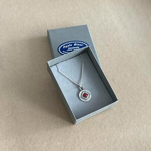 May include: A silver necklace with a circular pendant featuring a small red stone in a square setting, presented in a grey box with a blue label reading Ayala Binor Handmade silver jewellery. The pendant has a textured finish and a thin silver chain.