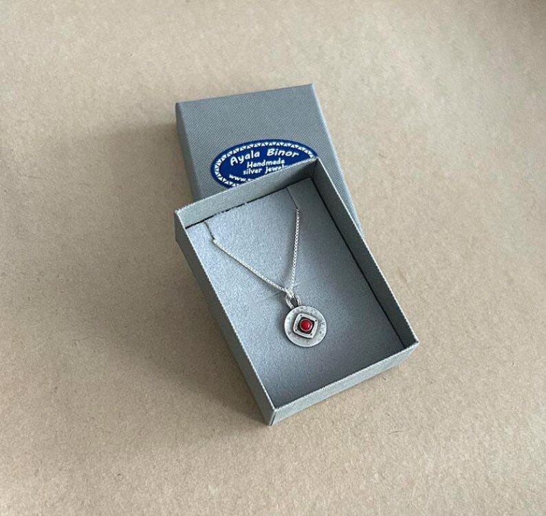 May include: A silver necklace with a circular pendant featuring a small red stone in a square setting, presented in a grey box with a blue label reading Ayala Binor Handmade silver jewellery. The pendant has a textured finish and a thin silver chain.