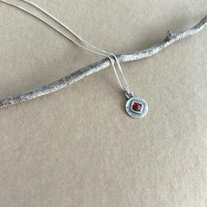 May include: A silver necklace featuring a small, circular pendant with a textured, brushed finish. The centre of the pendant contains a raised square frame holding a single, small, round, bright red stone. The chain is a thin, delicate silver link.