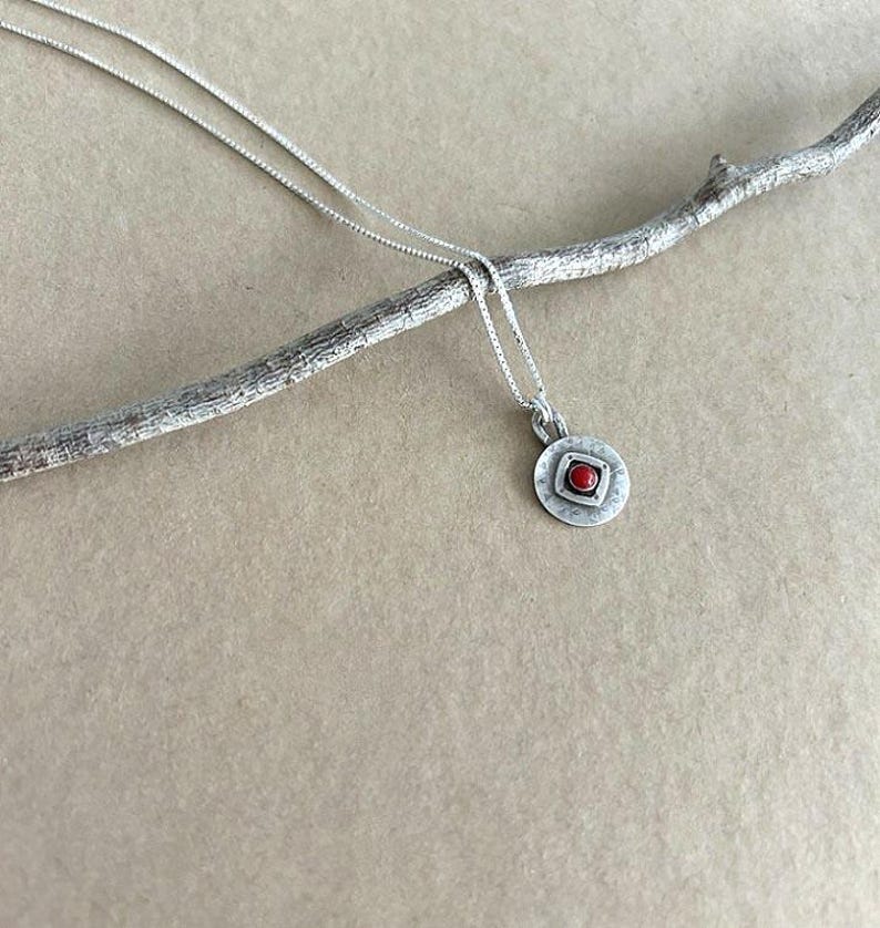 May include: A silver necklace featuring a small, circular pendant with a textured, brushed finish. The centre of the pendant contains a raised square frame holding a single, small, round, bright red stone. The chain is a thin, delicate silver link.