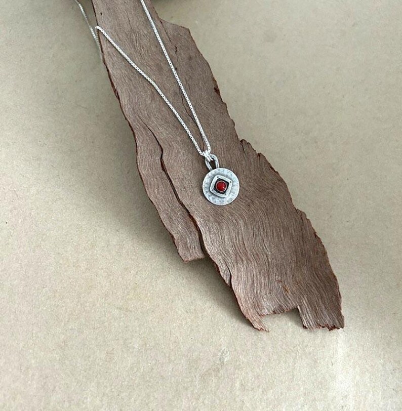May include: A silver box chain necklace featuring a circular pendant with a textured, hammered finish. The pendant contains a central square frame holding a small, round, vibrant red stone, creating a contrast between the metal and the gem.