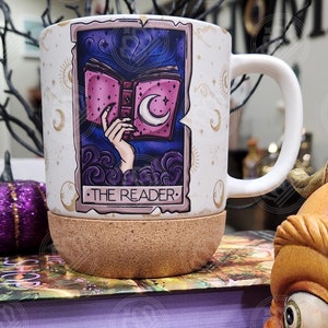 May include: A white ceramic mug with a cork bottom. The mug features a tarot card design with a hand holding a book with a crescent moon on the cover. The card is titled "The Reader".