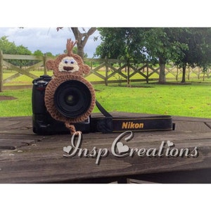 May include: A brown crocheted monkey camera lens cap with white eyes and a happy face. The lens cap is attached to a black camera with a Nikon strap.