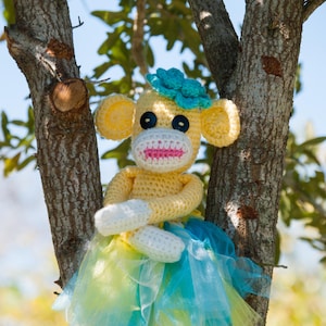May include: A crocheted monkey doll wearing a blue and yellow tulle skirt and a blue flower on its head. The doll is sitting on a tree branch.