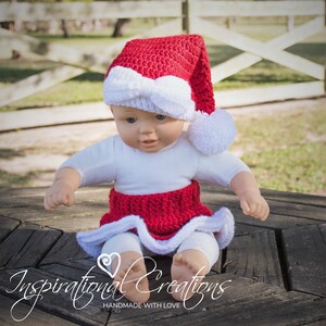 May include: A red and white crocheted Santa hat with a white pom-pom on top. The hat is worn by a doll sitting on a wooden surface.