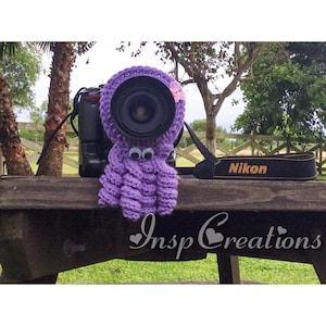May include: A purple crocheted octopus camera lens cover with googly eyes and a pink flower on a black camera with a Nikon strap.