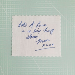 May include: A white fabric square with blue embroidered text that reads "Lots of love & a big hug from Gwen" with a line of blue embroidered "x"s below the signature.