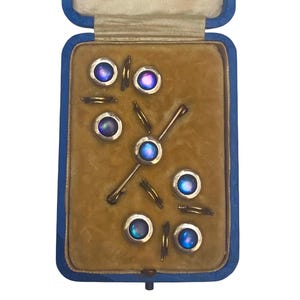 May include: A set of vintage iridescent cufflinks and shirt studs presented in a blue velvet-lined box. The cufflinks and studs feature a circular design with a central iridescent stone, set in gold-tone metal.