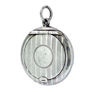 May include: A silver-coloured, round pendant with a hinged lid and a small loop at the top. The pendant's surface has vertical lines and a circular design in the centre. The pendant is likely a locket or a small container.