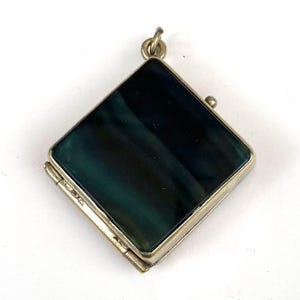 May include: A square-shaped locket pendant featuring a dark, marbled blue and black stone inlay set within a metallic frame. The piece includes a hinged side, a small circular bail at the top for attachment, and a tiny spherical knob on the right edge.
