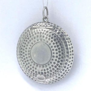 May include: A round, silver locket with a hammered texture. The locket has a hinged opening and a small loop for a chain. The front features a circular, raised design in the centre. The locket is suspended by a thin, silver wire.