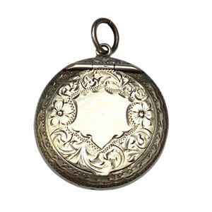 May include: A silver-coloured, round pendant with detailed floral and scrollwork engravings. The pendant has a hinged design and a small loop at the top for a chain. The centre has a blank, shield-shaped area. The overall design is ornate and detailed.