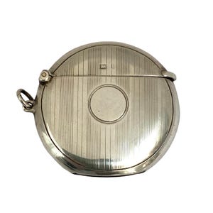 May include: A round, silver-coloured metal compact case featuring a hinged lid with a vertical pinstripe pattern and a central circular engraving area. The compact includes a small side loop for attachment and a visible hallmark on the top surface.