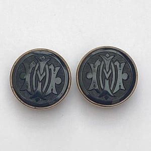 May include: Two dark grey buttons with a gold-coloured rim. Each button features a stylized monogram design in a lighter shade of grey. The buttons are round and appear to be made of a hard material.