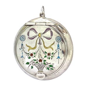 May include: A circular, silver pendant with a hinged lid. It features a painted design of a purple bow, gold ribbons, and a basket of flowers in red, green, and white. The pendant has a small loop at the top for a chain.