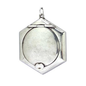 May include: A silver hexagonal locket with a circular space and a small loop for a chain. The locket has a hinged top and a heart-shaped clasp at the bottom.