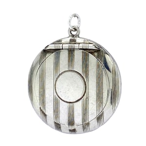 May include: A circular, silver-coloured locket with vertical striped detailing. The locket features a hinged opening and a small circular space in the centre. A small loop is at the top for attaching to a chain. The locket is likely vintage or antique.