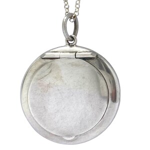 May include: A silver circular locket pendant with a hinged opening, suspended from a fine silver chain. The locket is open, showing a blank interior. The design is simple and elegant. The pendant is a classic piece of jewellery.