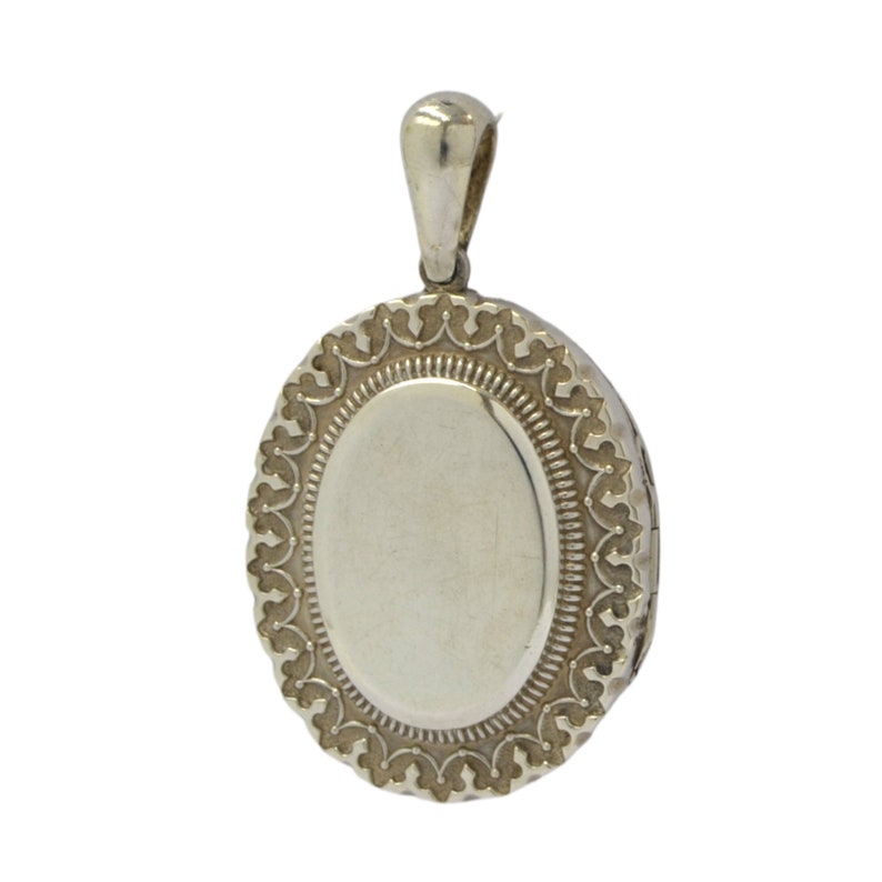 Op de afbeelding: Zilveren ovaal medaillon met een delicaat, versierd ontwerp rond de rand. Het medaillon heeft een scharnierend deksel en een lus om een ​​ketting aan te bevestigen.