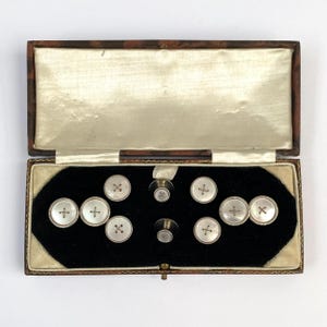 May include: A set of round, white mother-of-pearl shirt studs and cufflinks with silver-toned metal rims, each featuring a cross-stitch pattern, presented in a rectangular, hinged display box with a black velvet interior and cream satin lining.