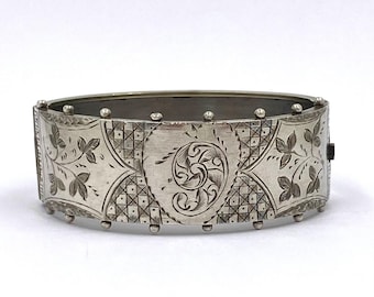 May include: A silver-toned hinged bangle bracelet. The bracelet features intricate engraved floral and geometric patterns. The central design is a swirling motif. The bracelet has a rectangular shape with rounded edges and small decorative studs.