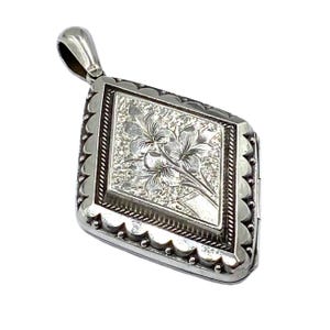 May include: A silver-toned, diamond-shaped locket with a floral design. The locket features a detailed, engraved floral pattern within a rectangular frame. The outer edges have a scalloped design, and a small loop is at the top for a chain.