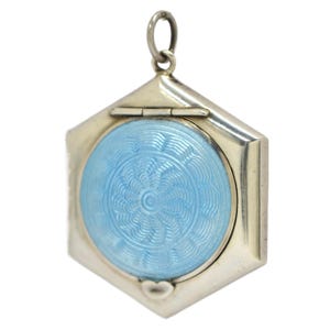 May include: A silver hexagonal pendant with a light blue, circular enamel centre. The enamel has a detailed, swirling pattern. A small heart shape is at the bottom. The pendant has a hinged top and a loop for a chain.