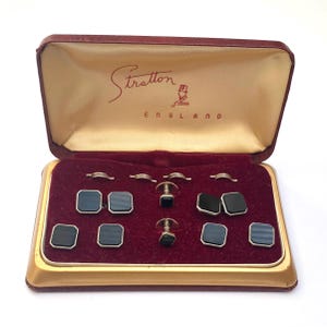 May include: A set of black square cufflinks and matching studs with gold-tone metal edges, presented in a maroon and gold-colored case with the text Stratton England printed on the interior lid above a small graphic of a person in a top hat.