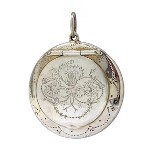 Peut inclure: Un pendentif médaillon rond en argent orné d'un motif de lyre gravé, entouré de volutes florales et de motifs de feuilles. La pièce métallique circulaire comprend une section supérieure articulée et un anneau de suspension en haut.