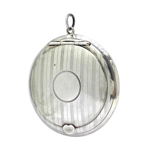 May include: A round, silver pendant with a hinged lid and a small loop at the top. The pendant features a striped pattern and a circular design in the centre. A heart-shaped detail is at the bottom.