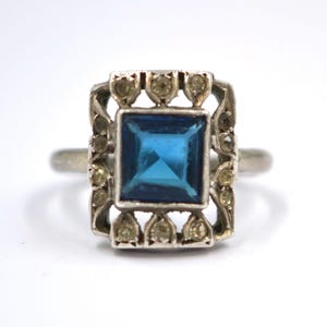 May include: A vintage silver ring featuring a square, vibrant blue gemstone at the center. The gemstone is surrounded by a decorative, openwork frame with smaller, light-colored stones. The ring has a classic, elegant design.