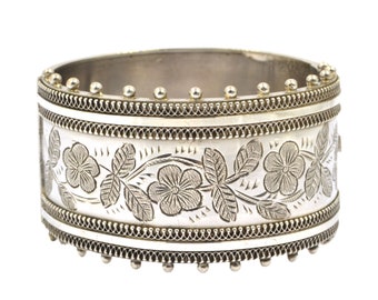 May include: A silver bangle bracelet with an engraved floral design. The bracelet has a beaded edge and a delicate floral pattern.