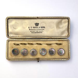 May include: A set of six circular buttons with iridescent mother-of-pearl centres and silver-toned rims, housed in a rectangular gold-trimmed case. The interior lid features the text E. T. BIGGS & SONS, Goldsmiths & Jewellers, UNDER THE CLOCK, MAIDENHEAD.