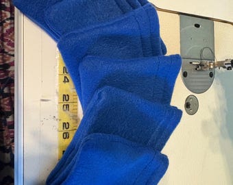 May include: Bright blue fleece fabric folded and stacked, possibly for a sewing project. The fabric is next to a measuring tape with inch markings. The fabric is on a white surface, near a sewing machine.