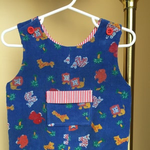May include: Blue corduroy overalls with a red and white striped pocket and a pattern of orange and red animals. The overalls have two red buttons on the straps.