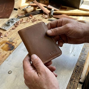 Puede incluir: Una cartera de cuero marrón con las iniciales "CRM" grabadas en la parte delantera. La cartera es rectangular con esquinas redondeadas y parece nueva. Las costuras son visibles alrededor de los bordes. La cartera está sujeta en las manos de una persona.