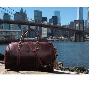 Puede incluir: Una bolsa de viaje de cuero marr&oacute;n con una correa para el hombro y detalles dorados. La bolsa est&aacute; sentada sobre una superficie de madera frente a una vista del puente de Brooklyn y el horizonte de la ciudad de Nueva York.