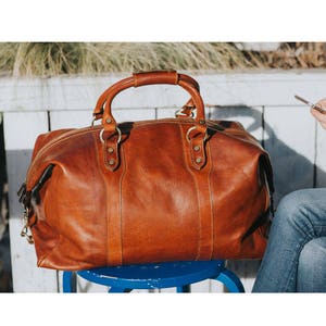 May include: A large, brown leather duffel bag with a top handle and a zipper closure. The bag has two smaller handles and metal hardware. It is sitting on a blue stool.