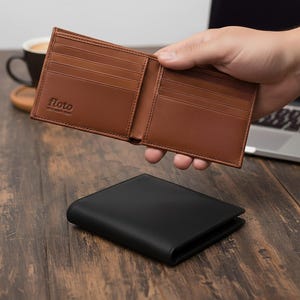 Puede incluir: Cartera de cuero marrón claro abierta con múltiples ranuras para tarjetas, sostenida sobre una cartera de cuero negro cerrada. La cartera abierta muestra la marca "floto". Las carteras están sobre una superficie de madera, con una taza de café y un portátil en el fondo.