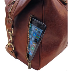 May include: Close-up of a brown leather duffel bag with a zippered pocket. A smartphone is partially visible inside the pocket. The bag has a gold-colored zipper and hardware. The brand name "floto" is visible on the zipper pull.