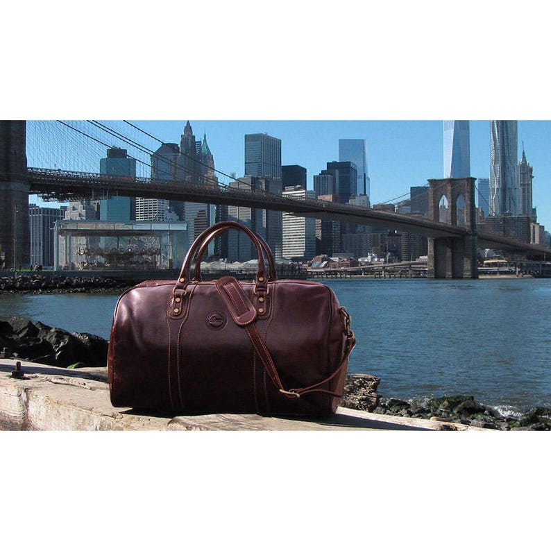 Puede incluir: Una bolsa de viaje de cuero marr&oacute;n con una correa para el hombro y detalles dorados. La bolsa est&aacute; sentada sobre una superficie de madera frente a una vista del puente de Brooklyn y el horizonte de la ciudad de Nueva York.