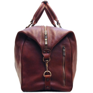 May include: A brown leather duffel bag with a top handle and a side zipper. The bag has a large main compartment with a zipper closure. The hardware is gold-colored. The bag is made of a smooth, high-quality leather.