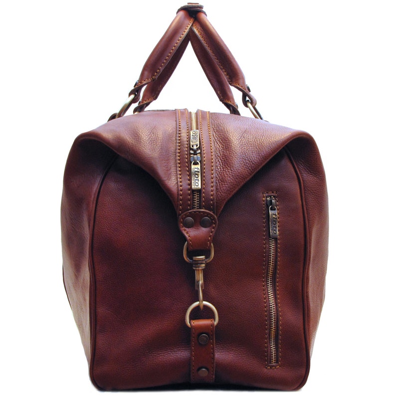 May include: A brown leather duffel bag with a top handle and a side zipper. The bag has a large main compartment with a zipper closure. The hardware is gold-colored. The bag is made of a smooth, high-quality leather.