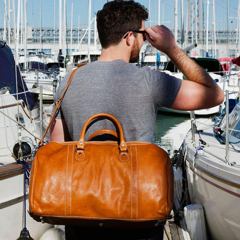 Puede incluir: Una bolsa de lona grande de cuero marr&oacute;n claro con una correa para el hombro y asas superiores. La bolsa tiene costuras visibles y herrajes met&aacute;licos. La imagen est&aacute; tomada al aire libre con veleros al fondo.