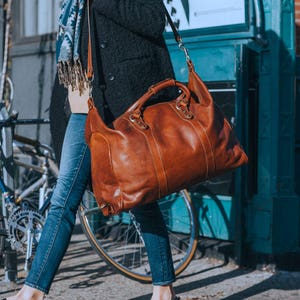 May include: A large, brown leather duffel bag with a top handle and a shoulder strap. The bag has a classic design with visible stitching and metal hardware. The bag is carried by a person wearing blue jeans and a black coat.