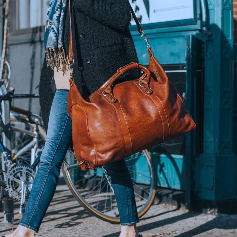 May include: A large, brown leather duffel bag with a top handle and a shoulder strap. The bag has a classic design with visible stitching and metal hardware. The bag is carried by a person wearing blue jeans and a black coat.