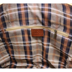 May include: Interior of a bag with a plaid pattern in shades of brown, tan, and navy blue. A gold zipper runs across the top, and a brown leather tag with the word "floto" is sewn inside.