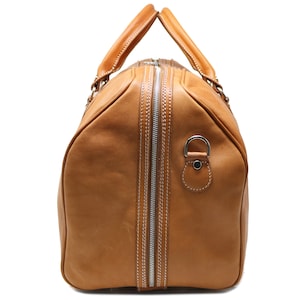 Pode incluir: Uma mala de viagem em couro castanho claro com um fecho de correr prateado no centro. A mala tem duas al&ccedil;as superiores e uma al&ccedil;a lateral. O couro &eacute; costurado com linha branca. Ideal para viagens.